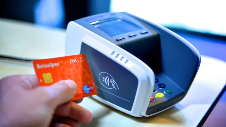 A hand holding a bank card beside a contactless card payment terminal